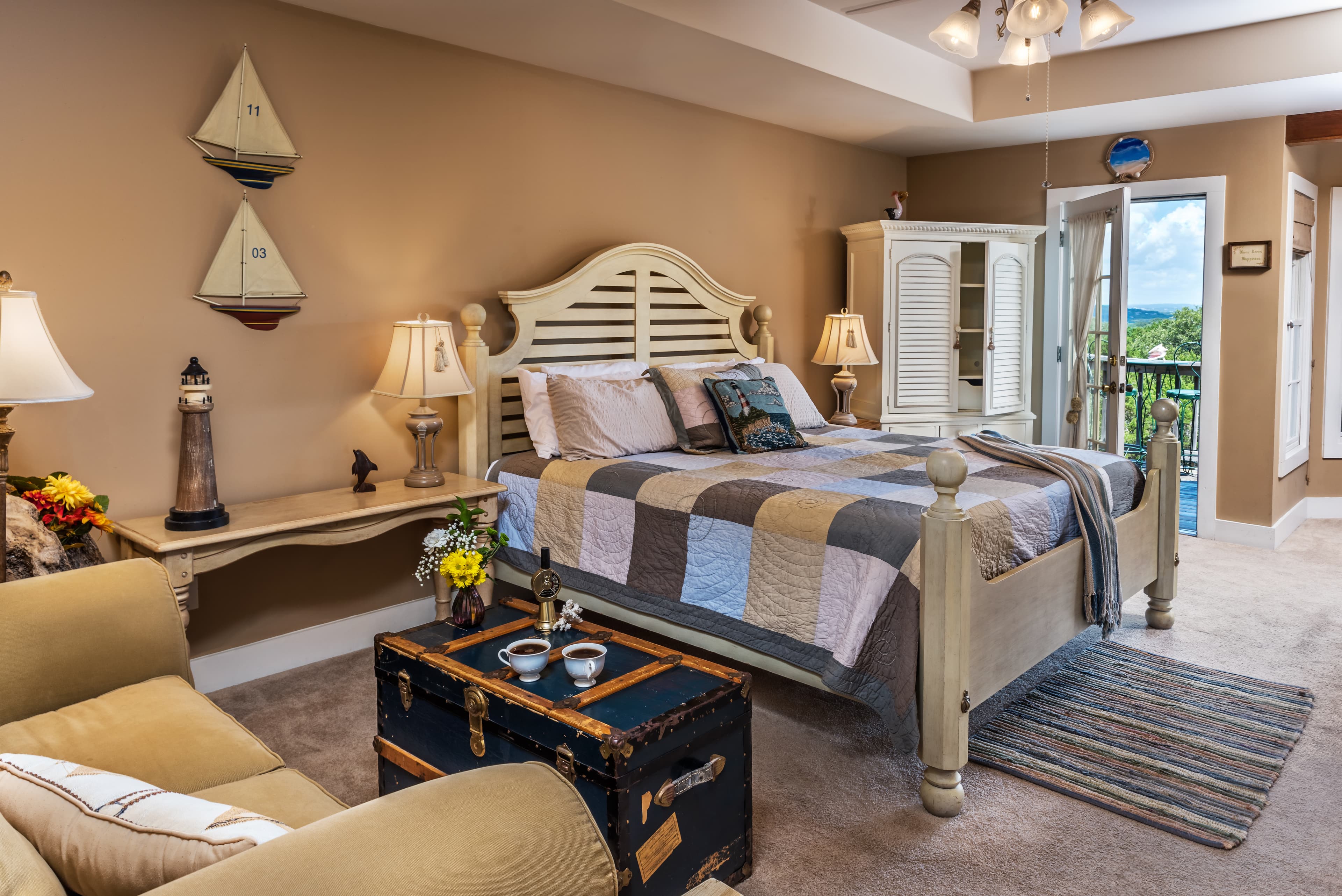 A bedroom decorated in a nautical theme. The bed has a patchwork quilt and there's an armchair nearby. There's a table with a vase of flowers, and a large window with a view of the outdoors.