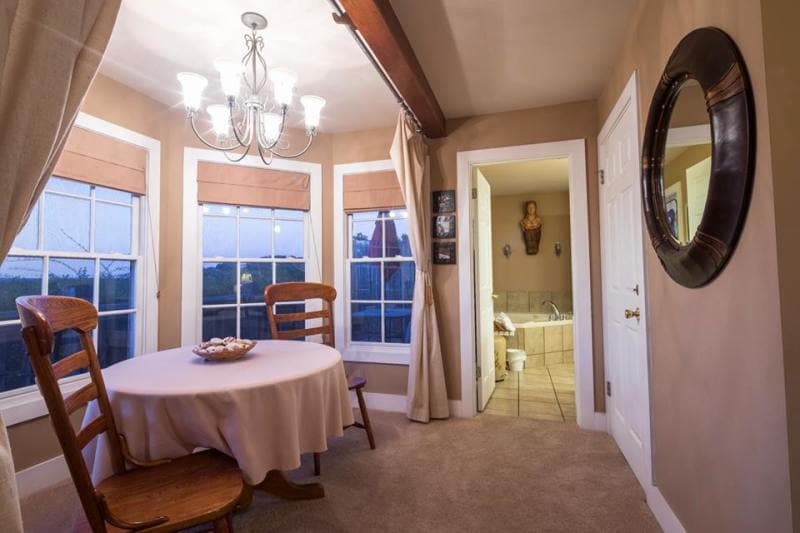 A small, cozy dining area with a round table and four chairs. The room has large windows with curtains and a view of the outdoors. A doorway leads to a bathroom with a bathtub visible.