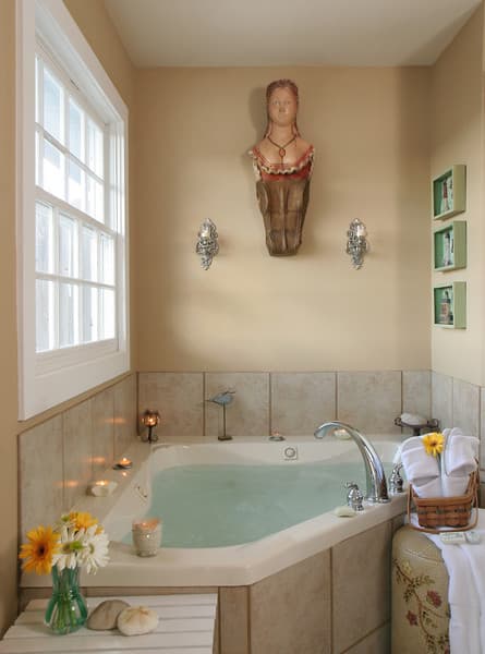 A spacious bathroom with a large, corner bathtub filled with water. There are candles on the ledge, flowers in a vase, and towels and a basket nearby. A figurehead from a ship's bow hangs on the wall.