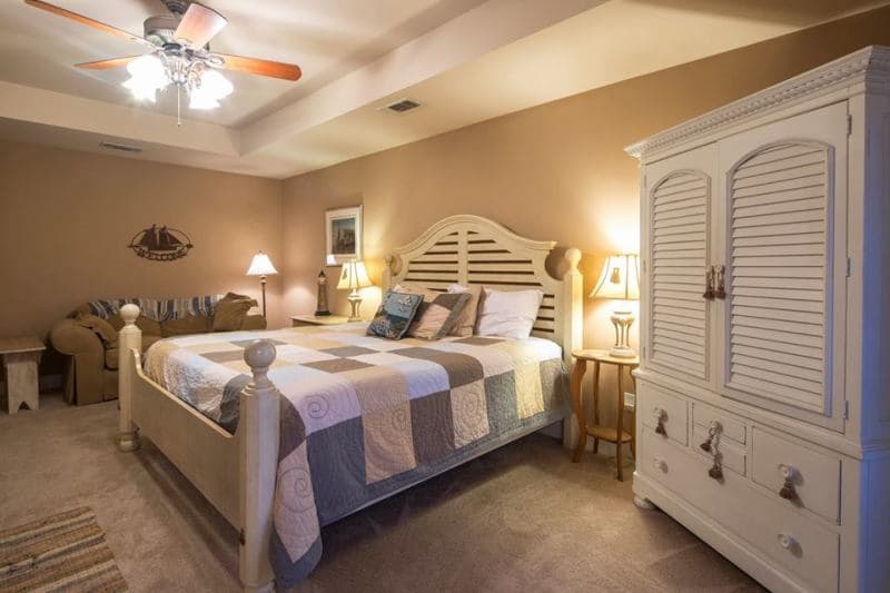 A large bedroom with a king-sized bed, a white wardrobe, and a small seating area with a couch and a table. The room has a ceiling fan and soft lighting.