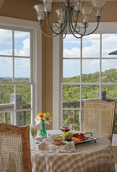 A table set for breakfast with a plate of fruit and pastries. The table is set with glasses of champagne and a vase of flowers. Large windows offer a view of a beautiful landscape with trees and hills.