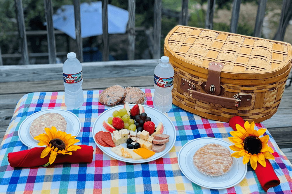 A picnic spread featuring plates of fruit, cheese, and meats, water bottles, and a wicker basket on a colorful checkered blanket.