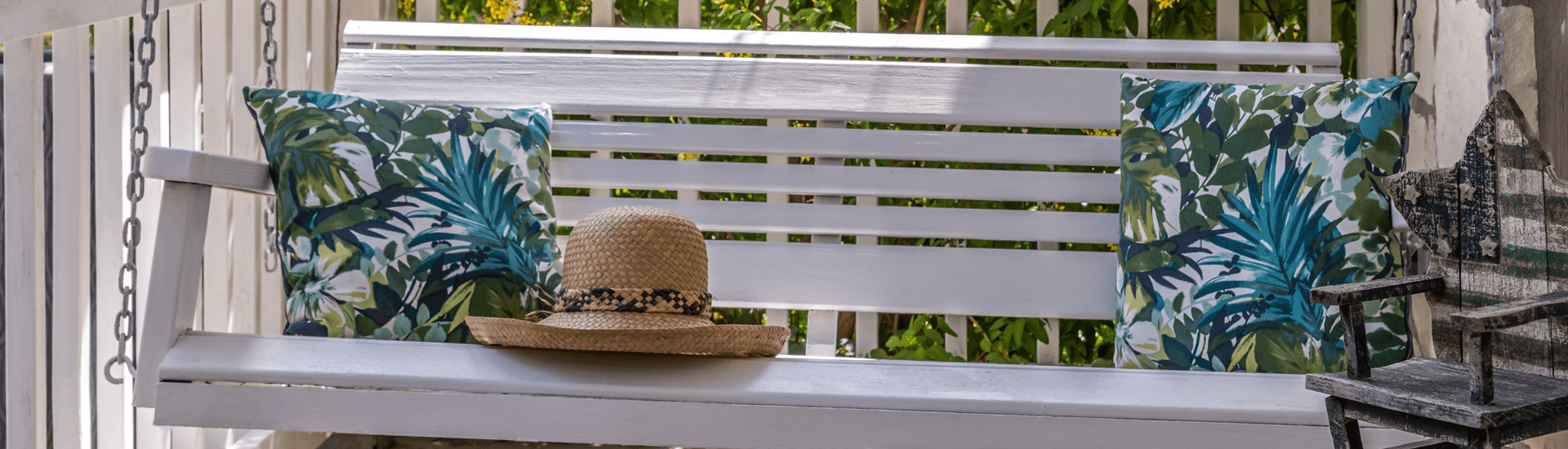 A white swing bench with tropical-patterned pillows and a straw hat resting on it.
