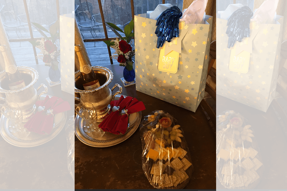 A festive table setting featuring champagne, decorative napkins, and a gift bag labeled "Baby Boy," accompanied by a flower arrangement and a platter of snacks.
