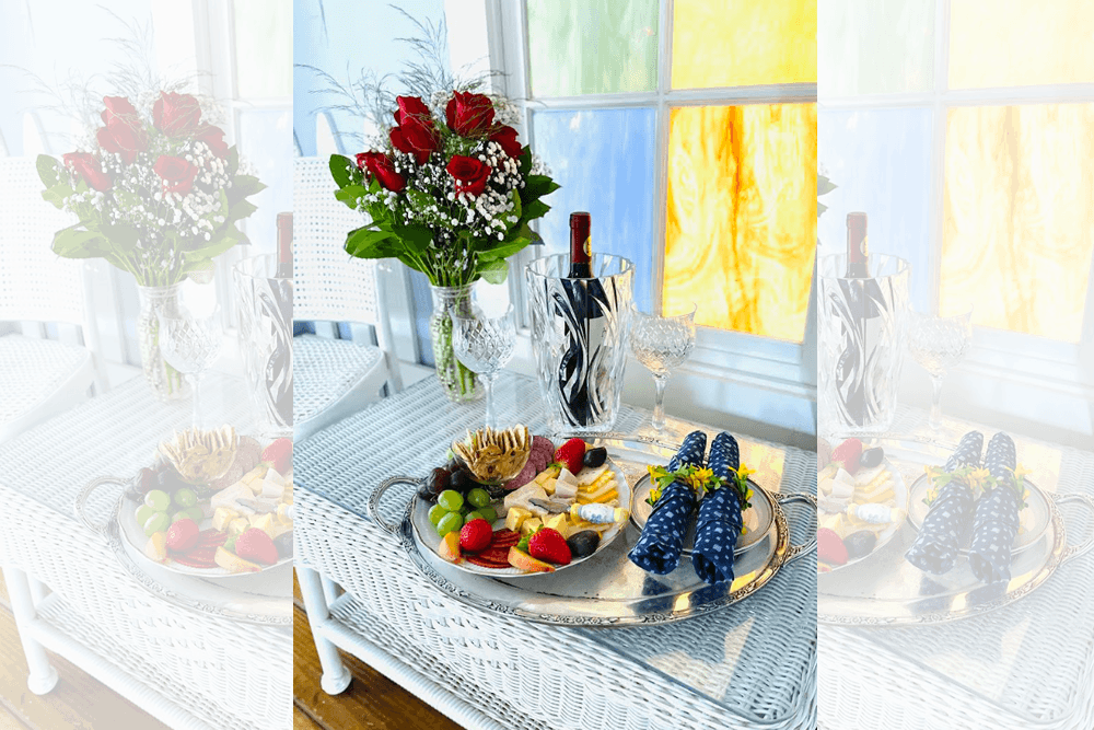 A cozy table setting featuring a bouquet of red roses, wine, and an array of fruits and cheeses on a silver platter.