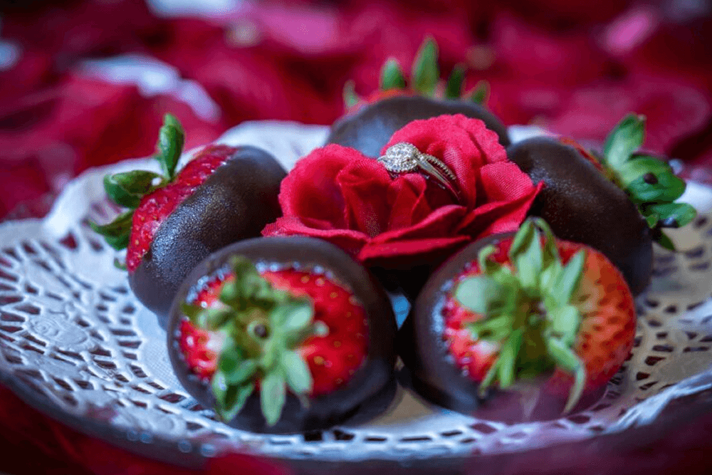 A plate of chocolate-covered strawberries garnished with a red rose and a diamond ring.
