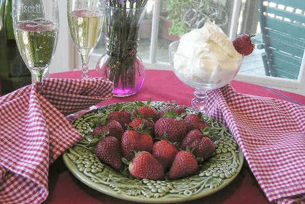 A plate of fresh strawberries surrounded by champagne flutes and a dessert bowl of whipped cream, all on a red tablecloth.