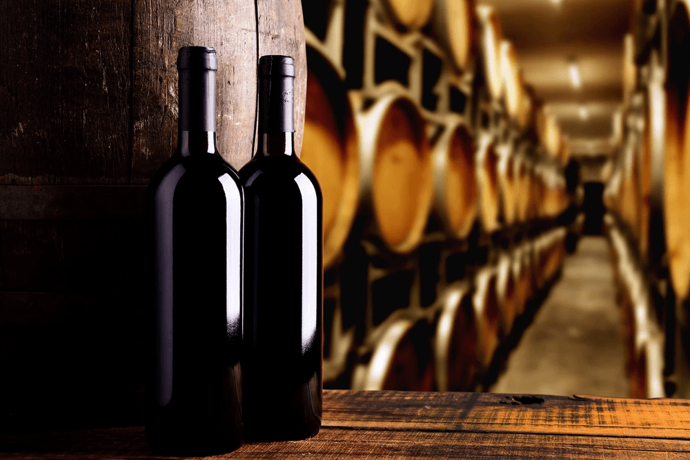 Two wine bottles in front of wooden barrels in a wine cellar.