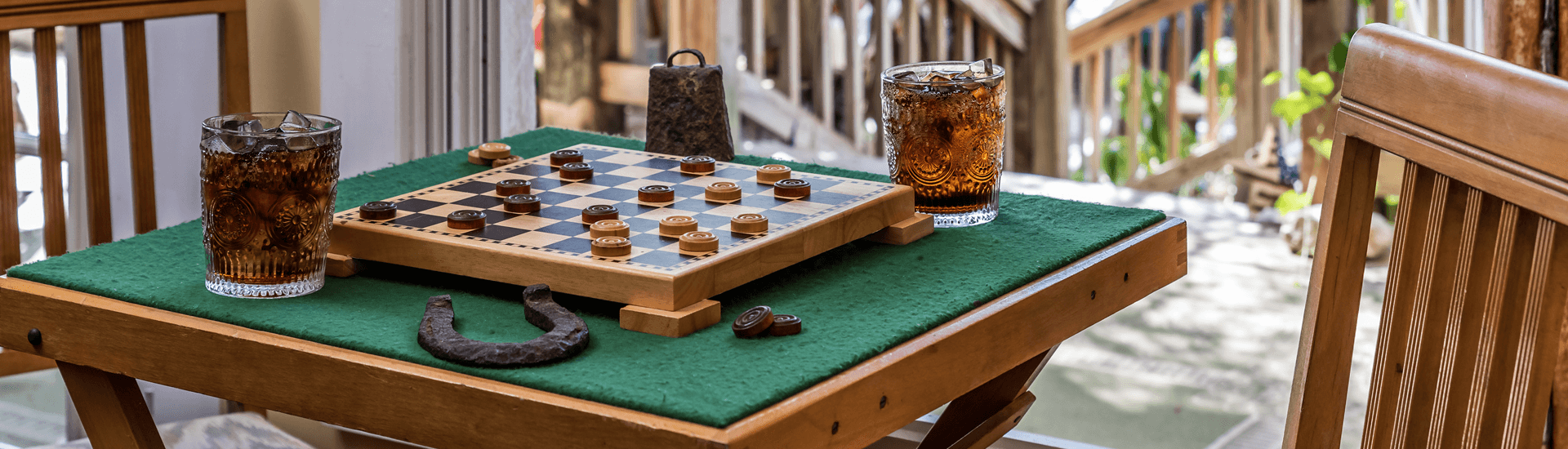 A wooden table with a green felt covering displays a checkers game, two glasses of iced tea, and a horseshoe.