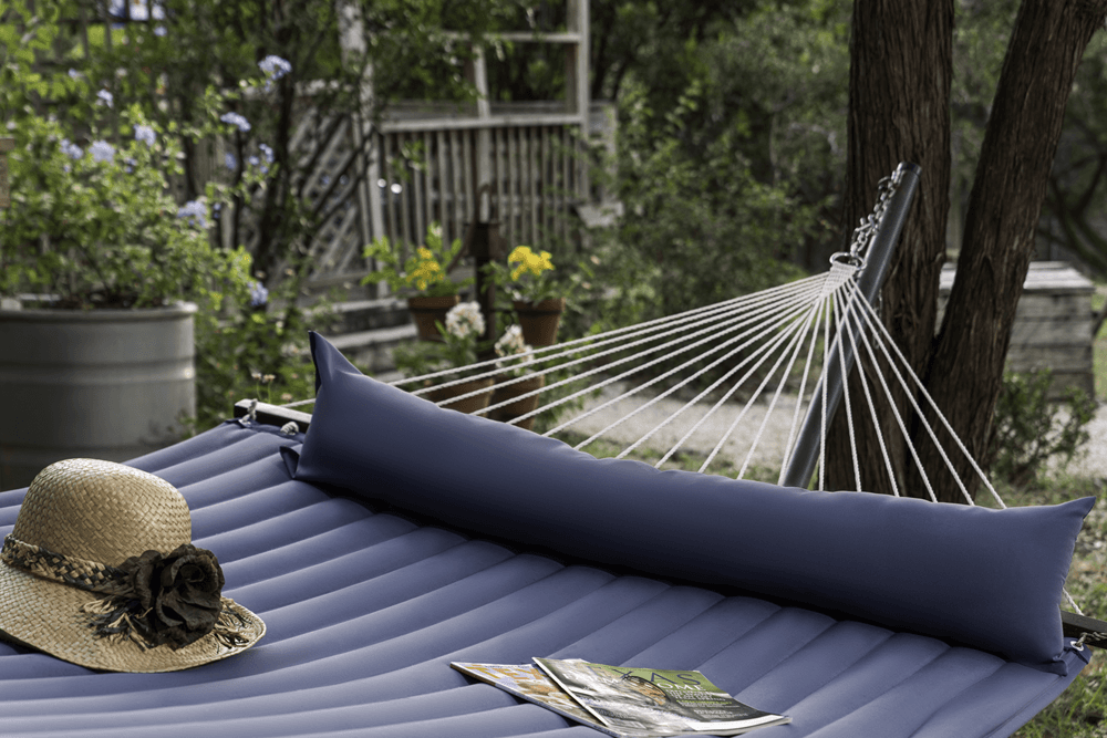 A straw hat and magazines rest on a blue hammock surrounded by greenery.