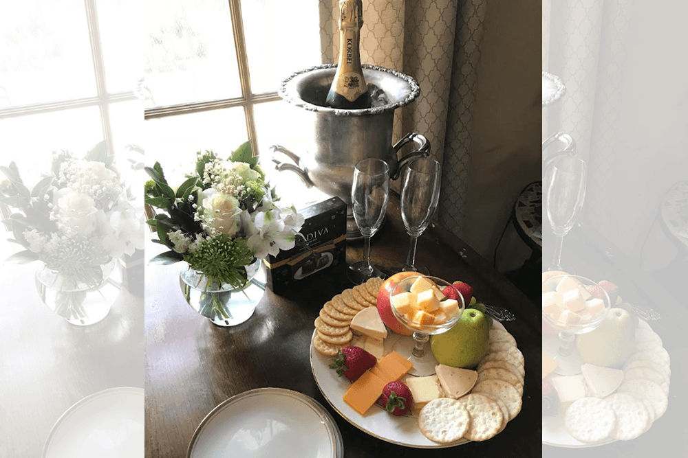 A table setting featuring a floral arrangement, champagne, and an assortment of cheeses, fruits, and crackers.