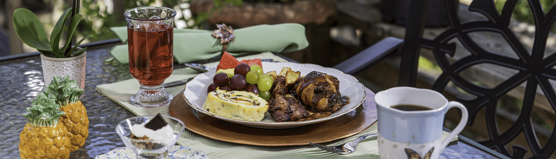 A beautifully arranged breakfast spread featuring an omelet, fruits, pastries, and drinks on a glass table with greenery in the background.