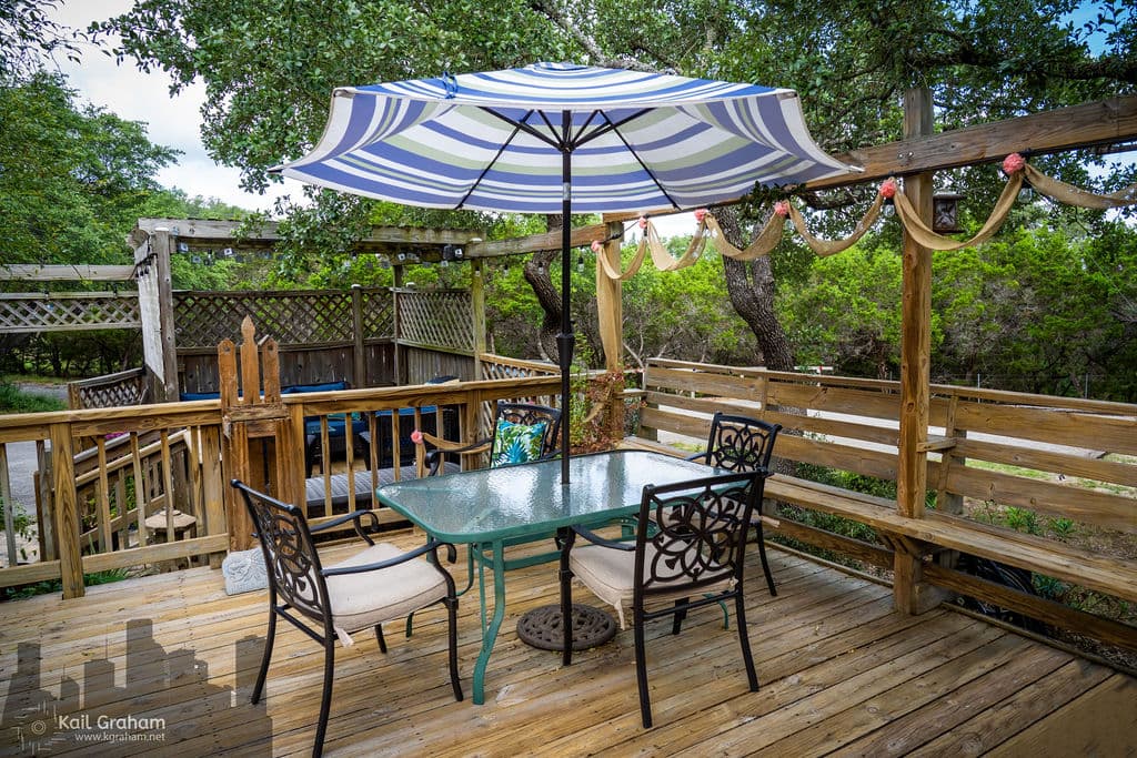 A wooden deck with a glass-topped table and chairs. A striped umbrella provides shade. There's a bench along one side of the deck, and a view of a backyard with trees and a fence.