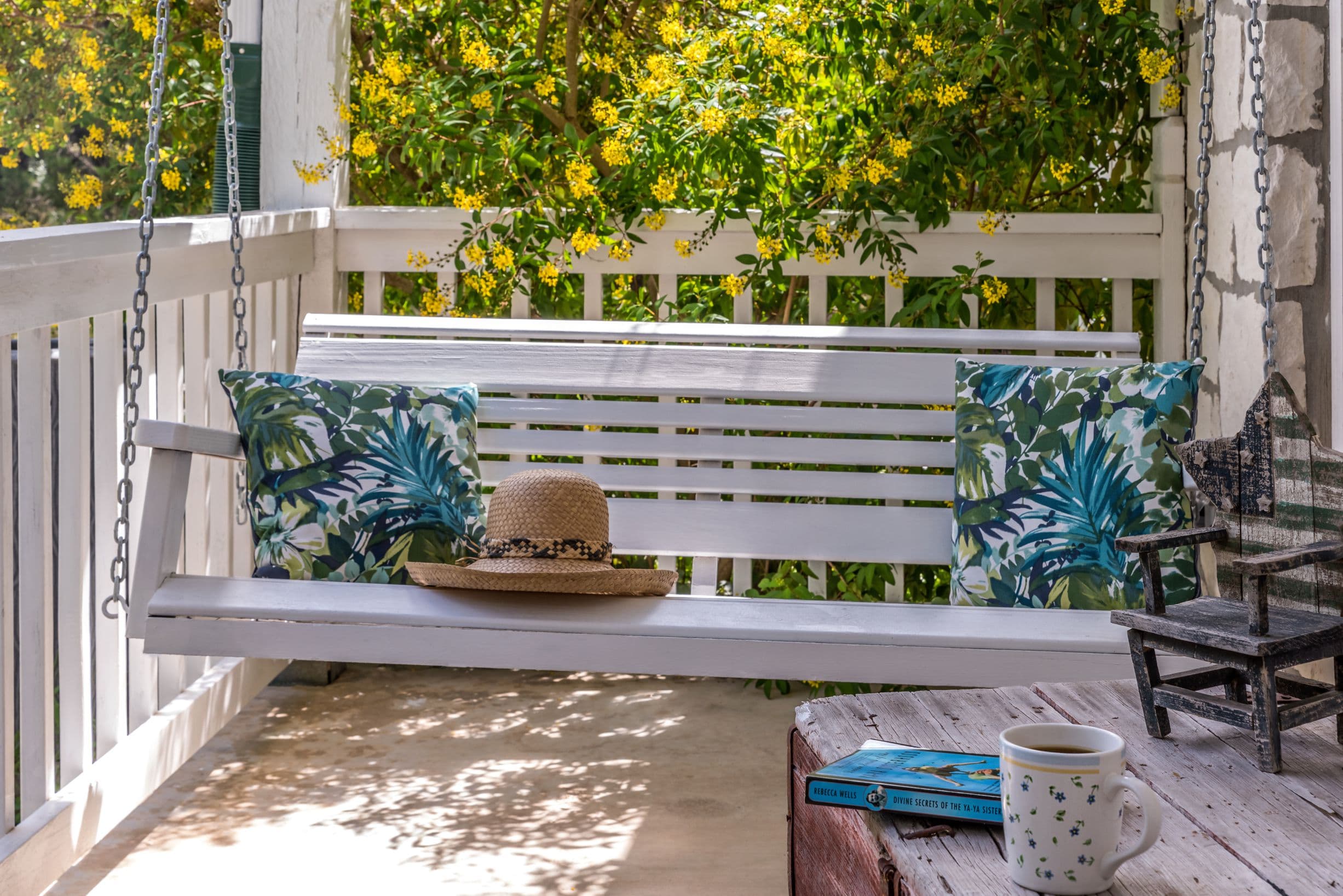 A white porch swing with colorful pillows and a sun hat resting on it. The swing is surrounded by lush greenery and a white picket fence.