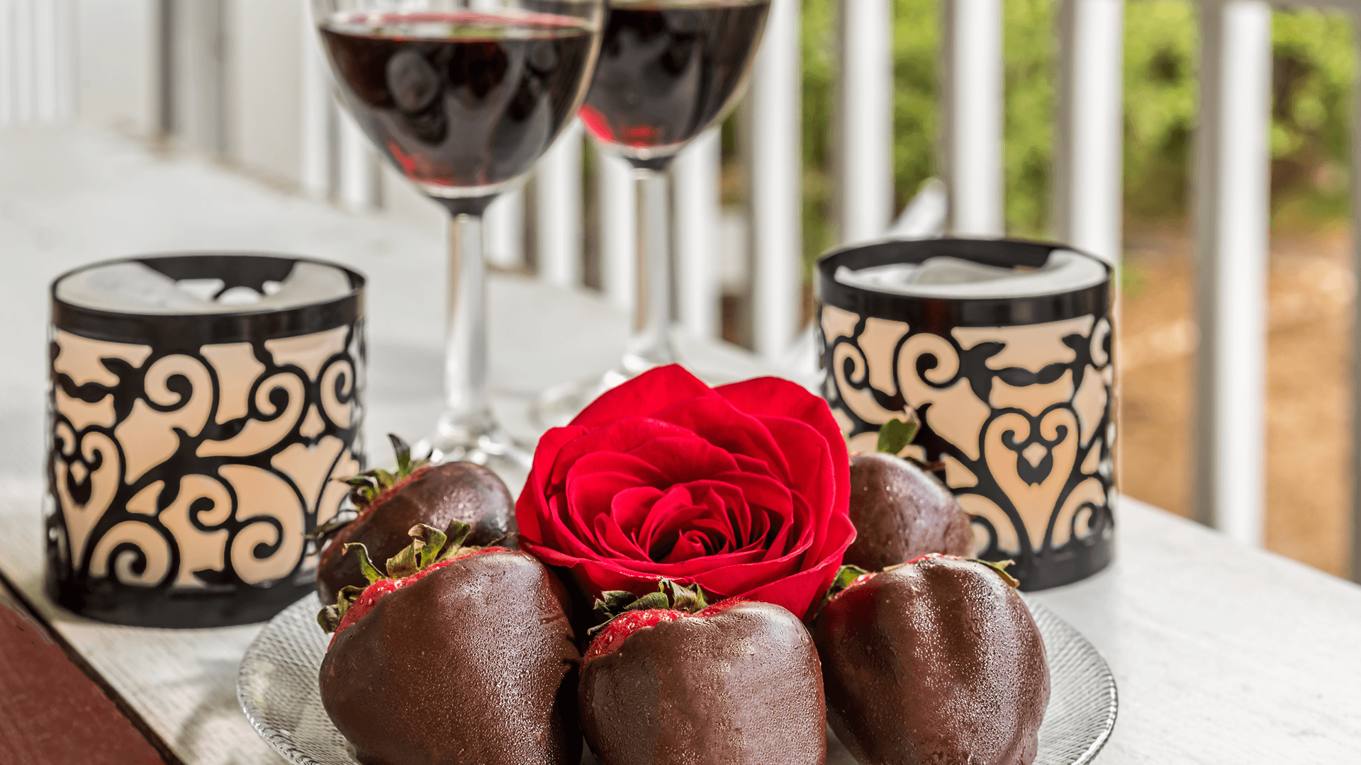 A romantic setup featuring chocolate-covered strawberries, a red rose, and two glasses of red wine beside decorative candles.