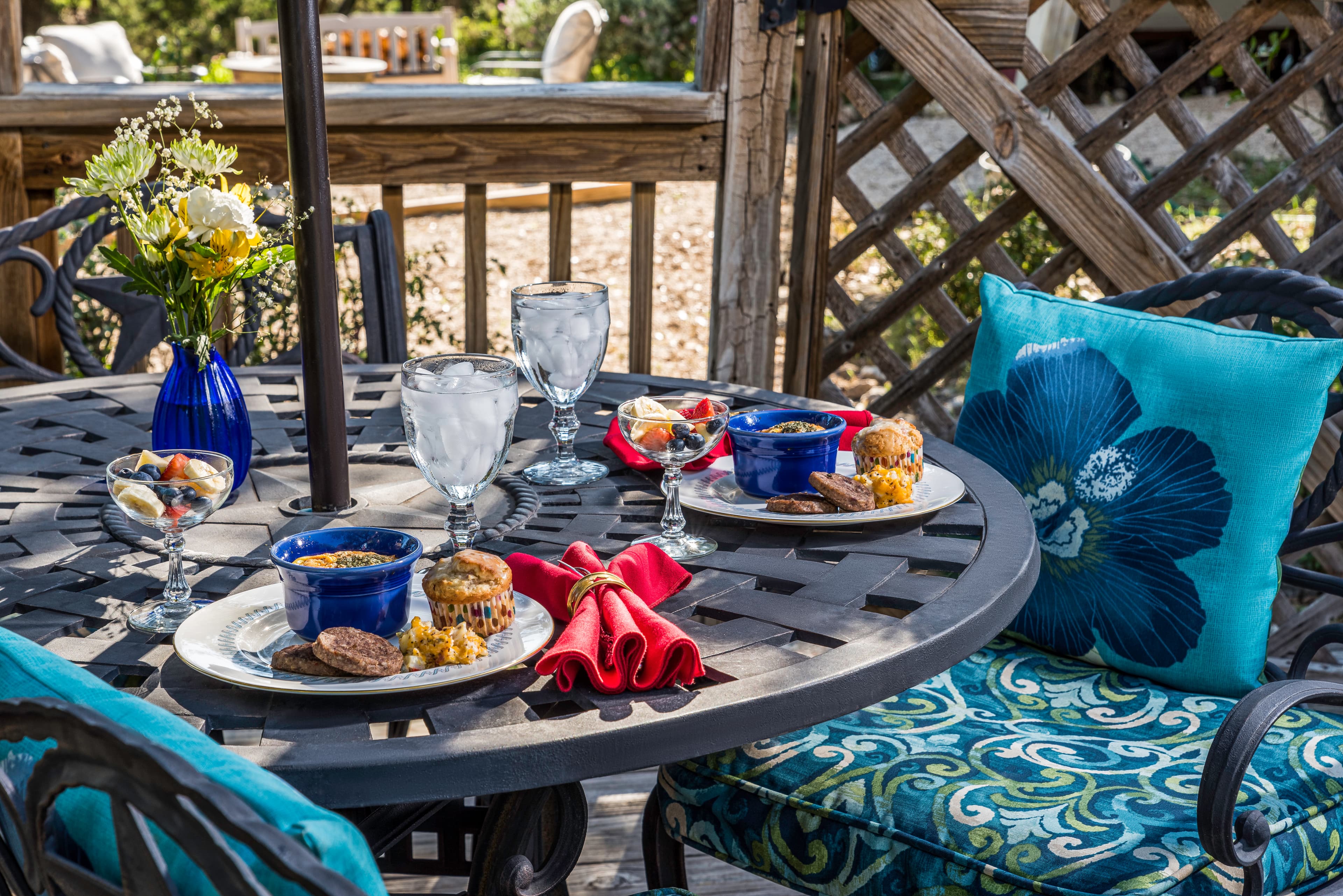 A beautifully set outdoor table with two plates of food, glasses of water, and decorative cushions.
