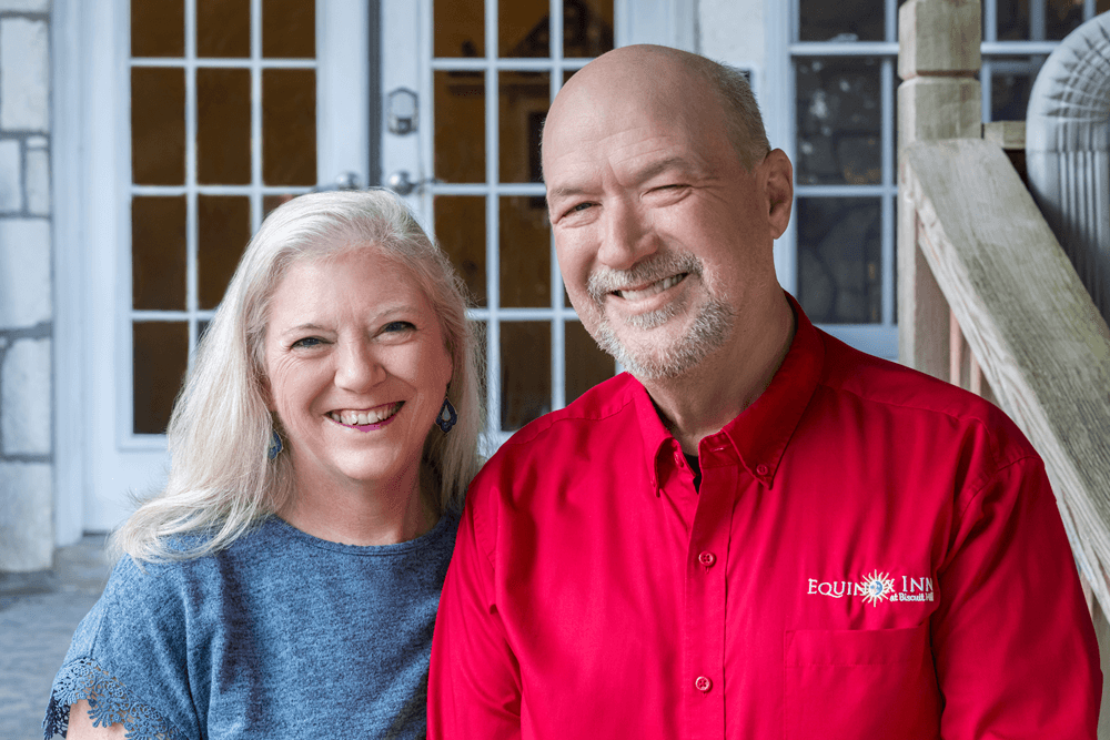 A smiling couple stands together on a porch, with a stone wall and glass doors in the background.