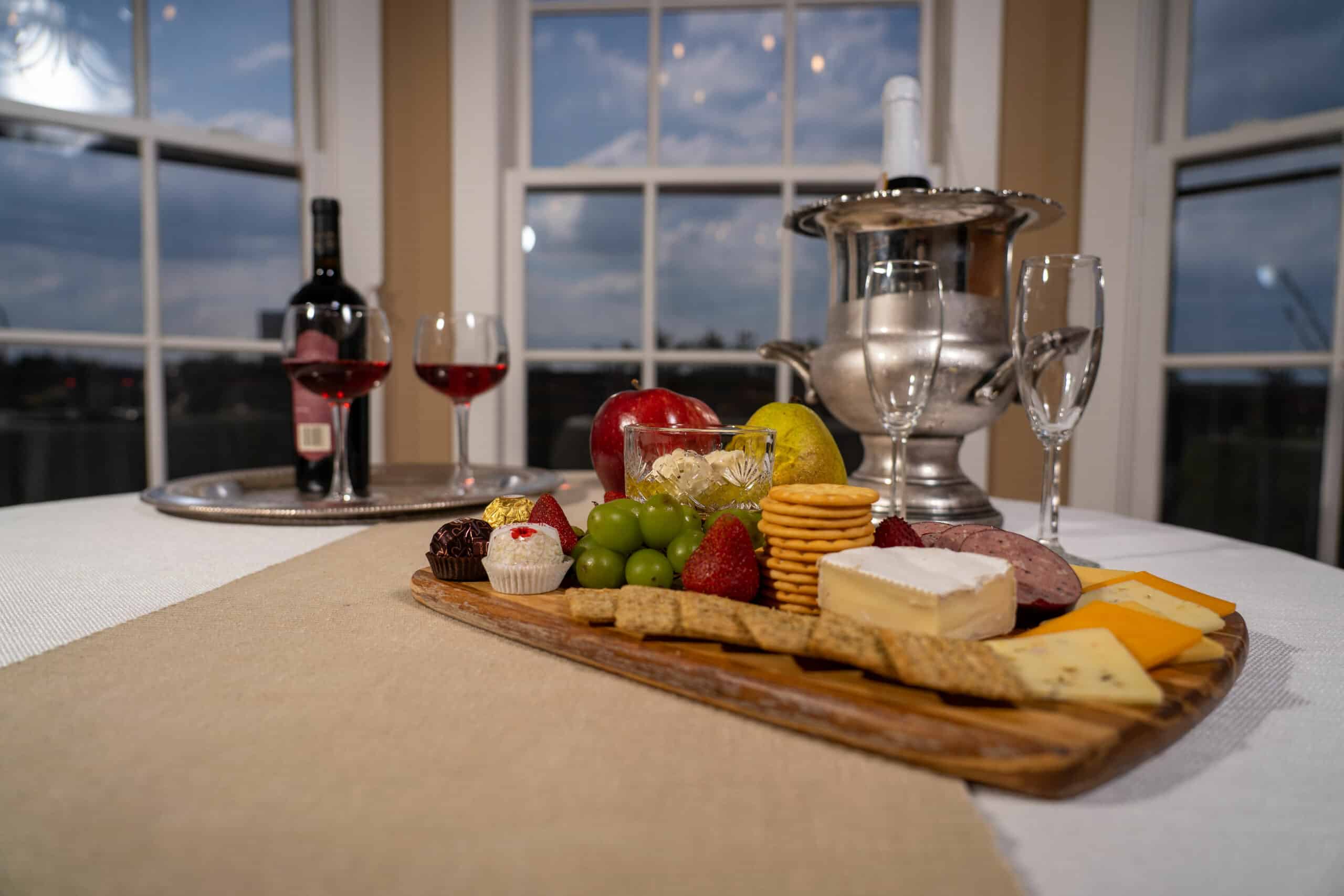 A beautifully arranged charcuterie board with various cheeses, fruits, chocolates, and a backdrop of wine and elegant glassware.