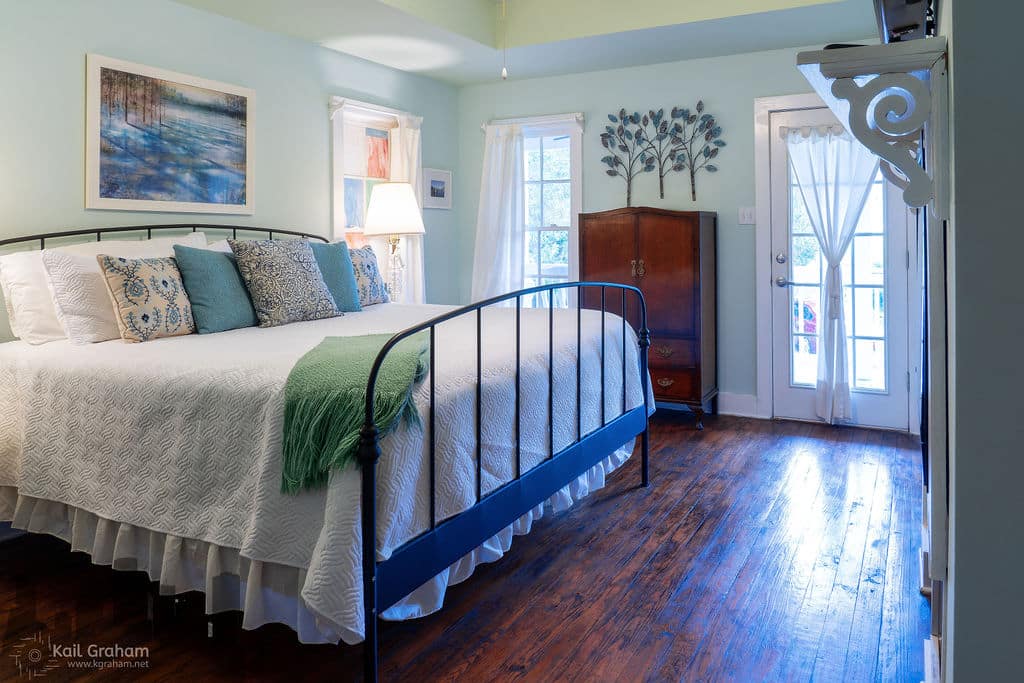 A well-decorated bedroom featuring a metal bed frame, colorful pillows, and a wooden dresser, with light streaming through a door and windows.