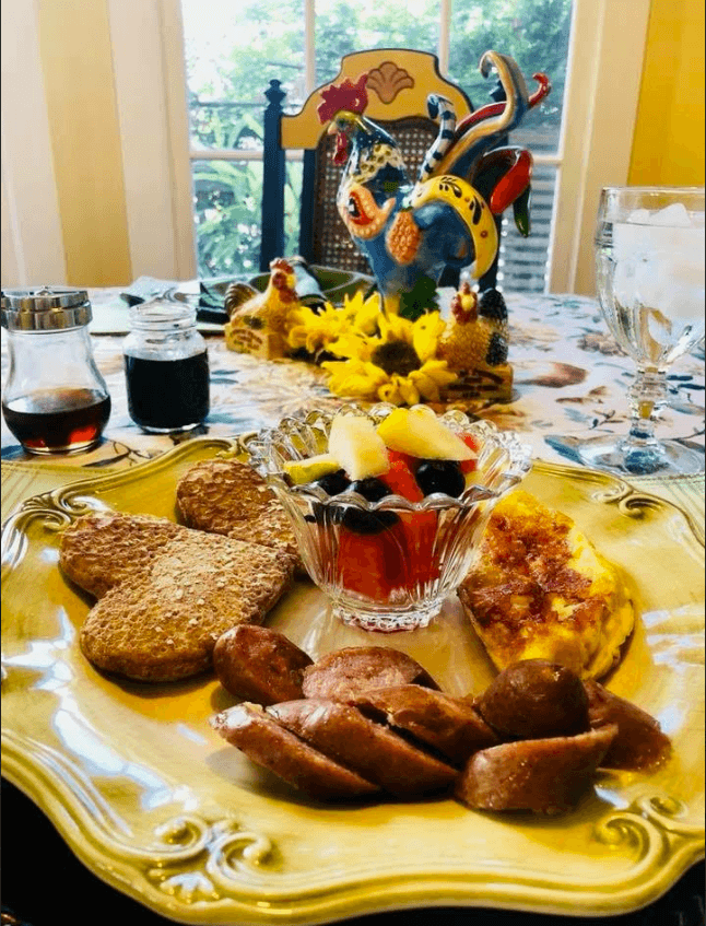 A beautifully arranged breakfast plate featuring fruit salad, sausage, pancakes, and baked goods, set on a vibrant table with decorative accents.