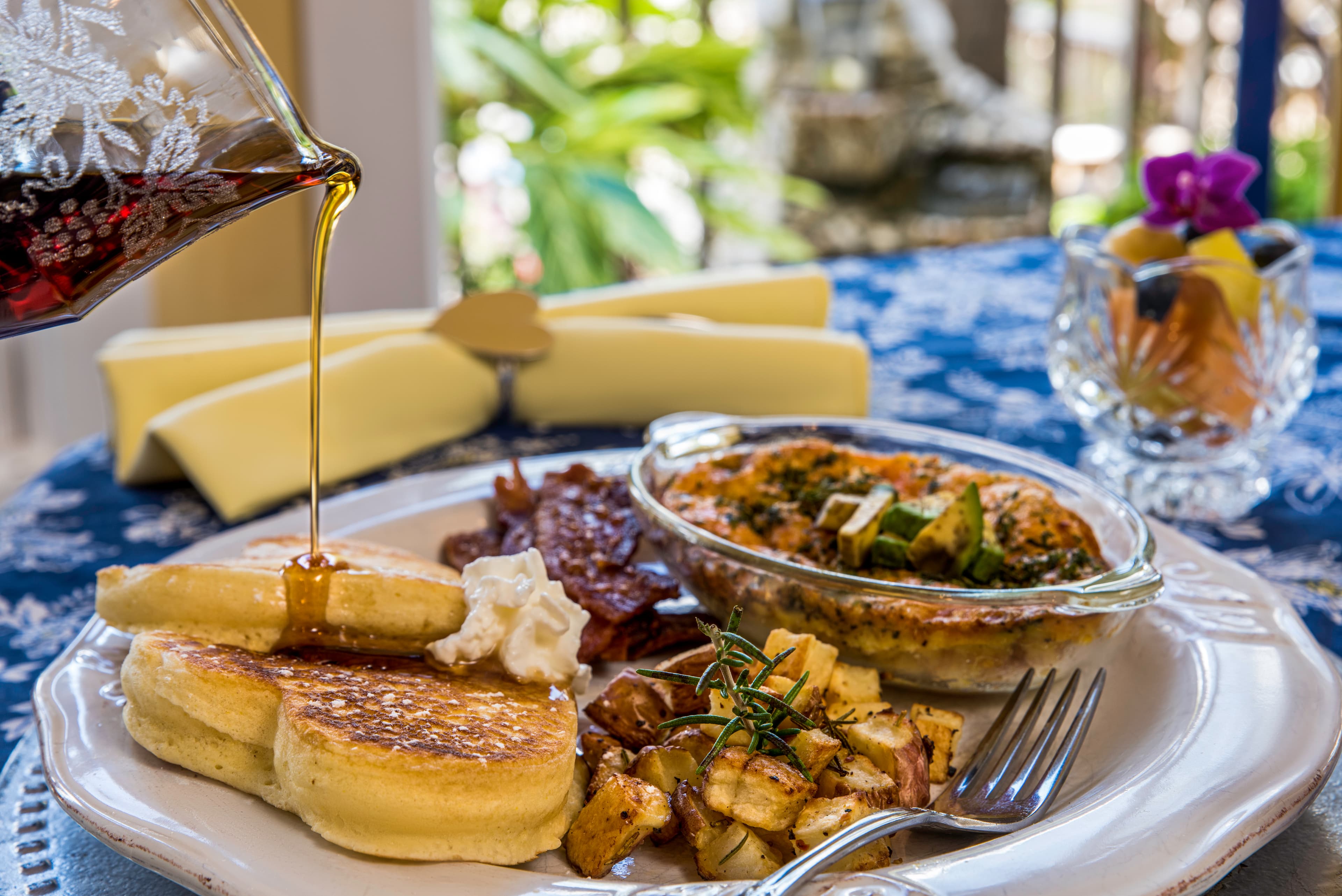 A plate of pancakes topped with syrup, accompanied by bacon, roasted potatoes, and a casserole.
